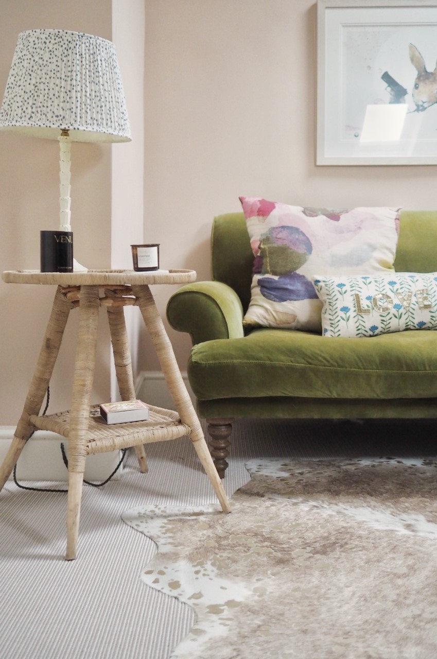 A setting plaster pink painted room with an olive green velvet sofa, round side table with blue and white spotty lamp shade. A neutral and gold spotty cow hide rug sits on the floor