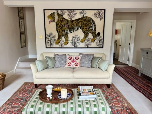 A living room with layered fabrics. The photo shows a large canvas tiger wall hanging with a cream sofa beneath with pretty scatter cushions. A large antique rug and a christopher farr cloth green ottoman