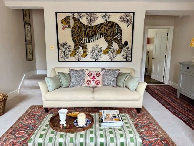 A living room with layered fabrics. The photo shows a large canvas tiger wall hanging with a cream sofa beneath with pretty scatter cushions. A large antique rug and a christopher farr cloth green ottoman